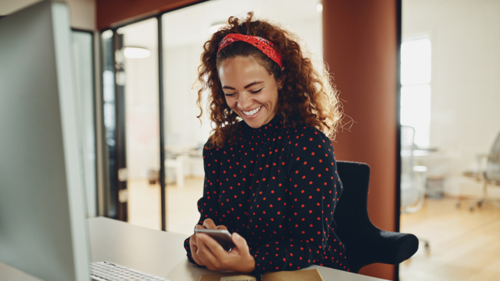 Frau sitzt am Schreibtisch und blickt lächelnd auf ihr Smartphone