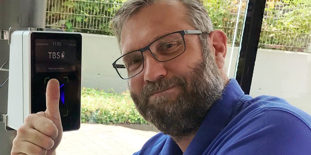 Interflex-Mitarbeiter mit biometrischem TBS-Terminal, blickt den Betrachter zufrieden mit "Daumen hoch"-Geste an