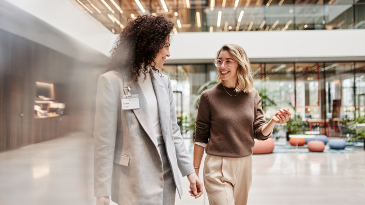 Zwei Frauen unterhalten sich lächelnd und laufen durch modernes Bürogebäude