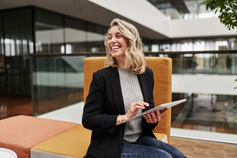 Frau sitzt lachend auf modernen Loungemöbeln und hält ein Tablet in den Händen